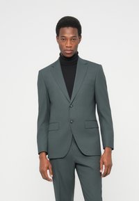 Ανδρικό κοστούμι σε σκούρο πράσινο ύφασμα, μονόπετο με δύο κουμπιά, πέτο με εγκοπή και εμπρόσθιες τσέπες. Φορεμένο πάνω από μαύρο ζιβάγκο.