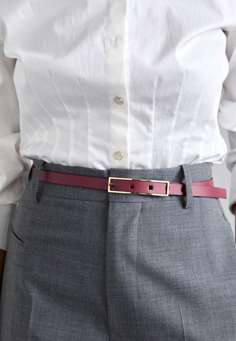 Fukszia öv sima bőrből, téglalap alakú arany csattal, szürke szabott nadrág és fehér ing felett viselve.