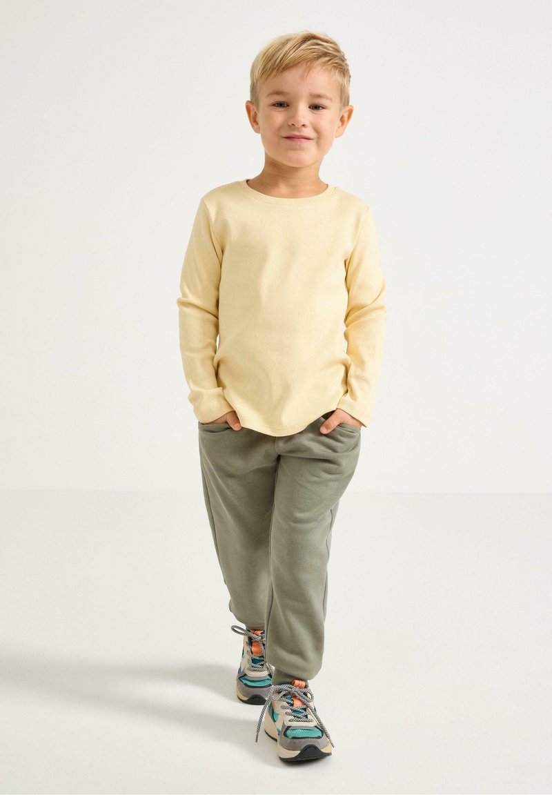 Geel shirt met lange mouwen en ribbelstructuur, gecombineerd met groene joggingbroeken. De outfit heeft sneakers met blauwe en oranje accenten.