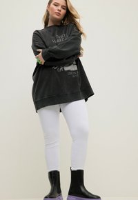 Ung kvinna med långt blont hår, iklädd en överstor svart grafisk sweatshirt, vita leggings och svarta stövlar med lila sulor, står med armarna i kors.