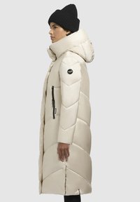 Lys beige dunfrakke med høj krave, geometrisk quiltet mønster, lynlåslomme og sort logo-patch. Båret med en sort strikket beanie.