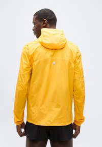 Άνδρας με π λάτη φοράει έντονο κίτρινο μπουφάν με κουκούλα και μαύρες βερμούδες πάνω σε απλό φωτεινό φόντο.