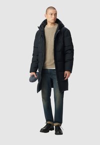 Cappotto imbottito nero con cappuccio, chiusura a zip frontale e tasche laterali, indossato sopra un maglione beige e jeans blu, completato con scarpe nere.