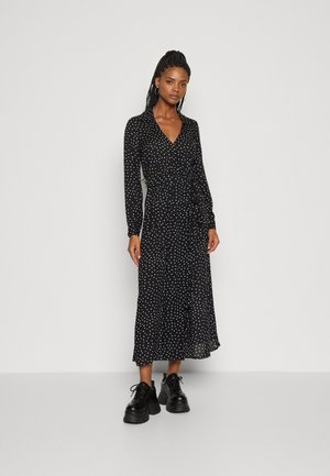 Robe longue noire à pois blancs, décolleté en V, manches longues, détail de taille nouée, tissu fluide, associée à des chaussures noires épaisses.