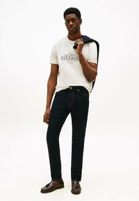 Slim-fit mørkeblå jeans parret med en lys t-shirt med et logo, båret med brune loafers og en navy sweater hængende over armen.