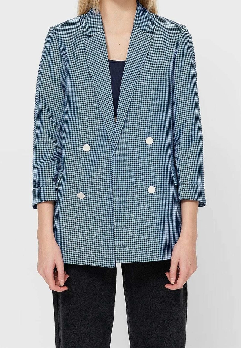 Femme portant un blazer croisé à carreaux bleu et blanc avec des boutons argentés et un pantalon noir sur un fond uni.
