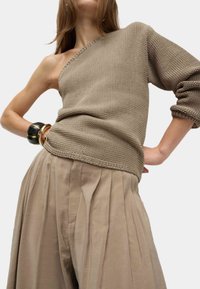 Pull en tricot beige asymétrique avec une manche longue, en tissu texturé, associé à un pantalon à pleats taille haute couleur tan.