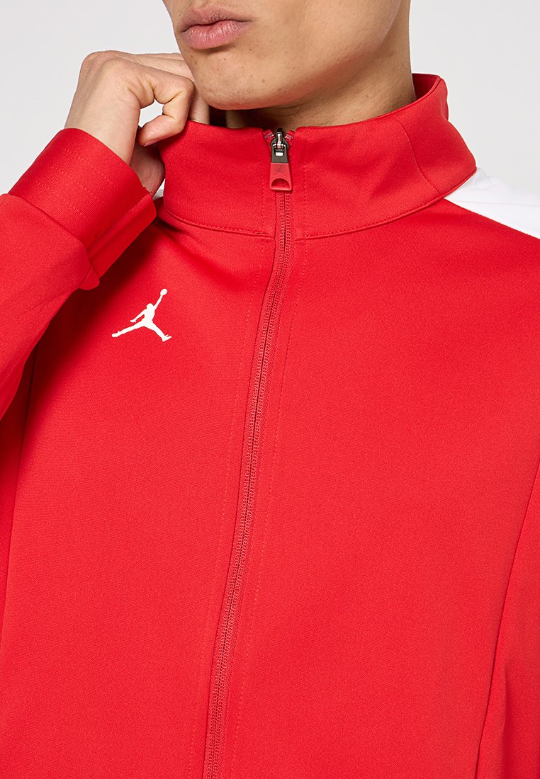 Chaqueta roja de cremallera con cuello alto, acentos blancos en los hombros y un pequeño logo Jumpman blanco en el lado izquierdo del pecho. Textura de tela suave.