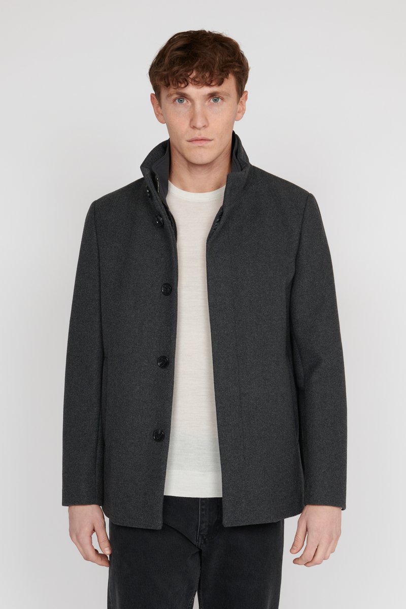 Grau wollener Mantel mit einem hohen Kragen, fünf großen schwarzen Knöpfen und einem geraden Schnitt, kombiniert mit einem hellen Pullover und dunklen Hosen.