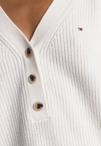 Detailní záběr na bílý pletený svetr ve tvaru V s třemi béžovými knoflíky a malým červeným, bílým a modrým logem Tommy Hilfiger.