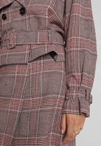 Manteau trench à carreaux rouges et noirs avec une taille ceinturée, un devant double boutonnage et un tissu texturé. Il présente des manches longues et des détails de boutons.