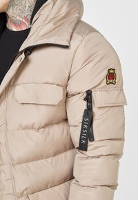 Giacca imbottita beige con collo alto, chiusura a zip e toppa sulla spalla. Dotata di tasca con zip con etichetta logo. Tessuto morbido e texturizzato.