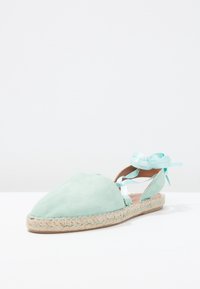 Espadrilles de ante en verde menta con punta redondeada, suela de yute y lazos de satén azul claro en el tobillo. Textura suave y lados abiertos.