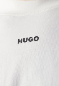 Valkoinen kangasmikä on ribbaustekniikalla, jossa on musta "HUGO" -logo keskitettynä rinnassa. Yksinkertainen pyöreä kaula-aukko, ei muita grafiikoita.