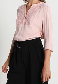 Blouse rose avec un panneau en dentelle au niveau de l'encolure, associée à un pantalon noir taille haute avec ceinture et plis.