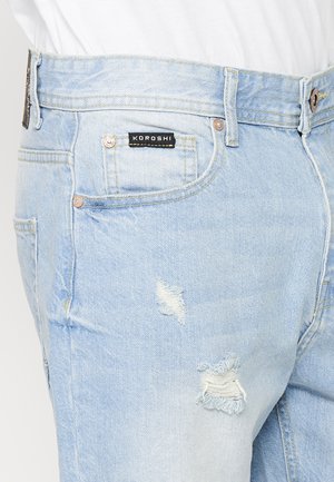 Lichtblauwe denim jeans met een verouderd ontwerp en gescheurde delen. Kenmerken zijn een zwart label met "KOROSHI" op de tailleband.