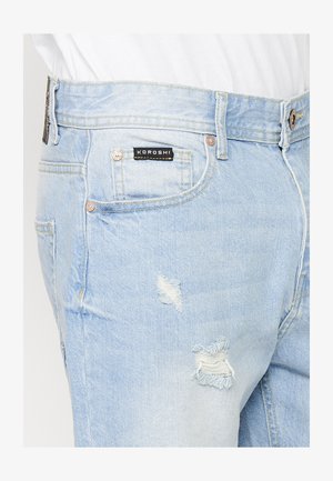 Helle blaue Jeans aus Denim mit einem lädierten Design und ausgefransten Rissen. Details umfassen ein schwarzes Label mit "KOROSHI" am Bund.