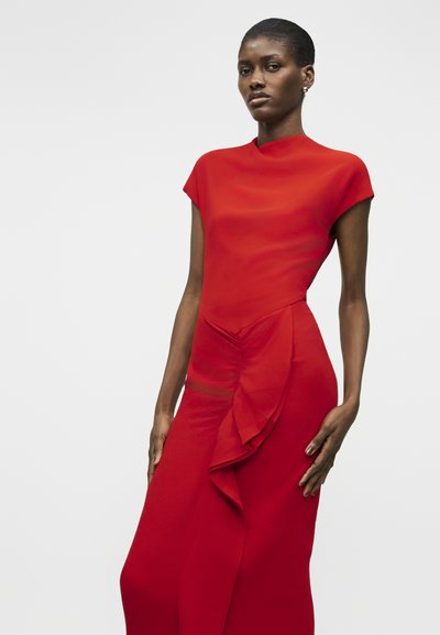 Mujer con un vestido rojo ajustado de manga corta y un detalle de volantes en la parte delantera, posando frente a un fondo claro y liso.