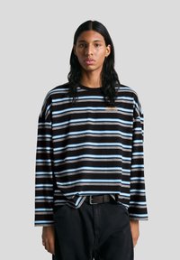 Fekete, hosszú ujjú csíkos ing kék és fehér csíkokkal, laza fazonnal és apró hímzett logóval a mellkason. Sötét nadrággal viselve.