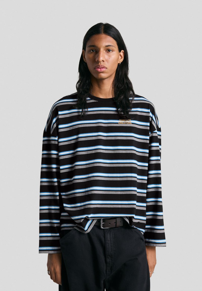 Fekete, hosszú ujjú csíkos ing kék és fehér csíkokkal, laza fazonnal és apró hímzett logóval a mellkason. Sötét nadrággal viselve.