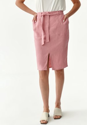 Vrouw draagt een knielange roze rok met een voorplooi, zakken en een ceintuur gestrikt in de taille, gecombineerd met witte open-toe sandalen met hak.