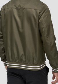 Oliwkowa kurtka bomber z gładką teksturą, ściągaczami na rękawach i białym pasek na dolnej krawędzi. Posiada lekko strukturalny design tylnej części.