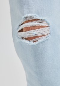Tissu en denim bleu clair avec une zone de genou usée présentant des bords effilochés et des découpes irrégulières pour un aspect texturé.