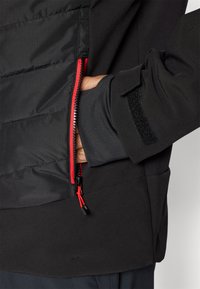 Schwarze Jacke mit einer Reißverschlusstasche mit rotem Reißverschluss, Klettverschluss am Ärmel und einer weichen, strukturierten Optik des Stoffes.