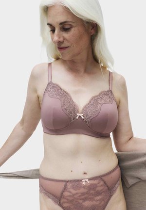 Frau mit heller Haut und langem weißen Haar trägt einen mauvefarbenen Spitzen-BH und passende Slip, hält einen beigen Stoff hinter dem Rücken.