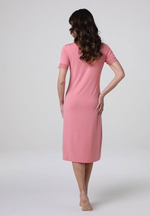 Donna con capelli castani ondulati, indossa un vestito rosa al ginocchio con maniche corte rifinite in pizzo, in piedi a piedi nudi di spalle davanti a uno sfondo semplice.