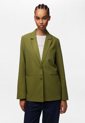 Giovane con capelli intrecciati che indossa una giacca verde oliva con bottoni sopra una maglia chiara e pantaloni scuri su uno sfondo bianco semplice.