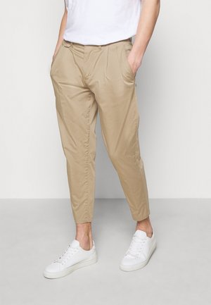 Homme portant un pantalon beige fuselé avec des poches latérales, associé à des baskets blanches et un t-shirt blanc, debout devant un fond uni.