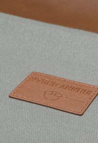 Tessuto grigio con una texture liscia presenta un'etichetta in pelle marrone impressa con "JOHNNY URBAN". L'etichetta ha bordi arrotondati e cuciture ordinate.