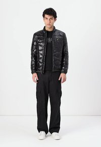 Giacca a vento nera con texture trapuntata, cerniera frontale e collo alto, indossata sopra una maglietta nera e abbinata a pantaloni cargo neri e sneaker bianche.