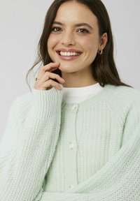 Cardigan en maille texturée vert clair avec un col rond et une fermeture boutonnée, doté de manches longues et d'une apparence douce et confortable.