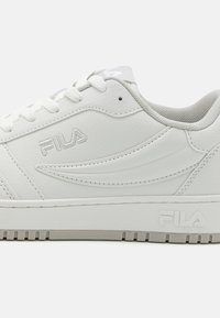 Vit lädersneaker med perforerade detaljer, platta snören och en strukturerad gummisula. Har en subtilt varumärkeslogotyp i grått på sidan.