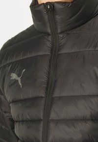 Crna puf jakna s visokim ovratnikom, prednjim patentnim zatvaračem i toniranim Puma logom. Ima štuckani uzorak i glatku površinu tkanine.