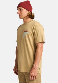 Beige bomull t-skjorte med rund hals, med et flerfarget grafisk design på brystet. Teksturert rød strikket lue båret på hodet.