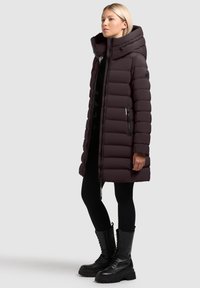 Parka in un profondo bordeaux con colletto alto e tasche con zip. Presenta un design trapuntato e maniche aderenti. Indossata sopra leggings neri e stivali.