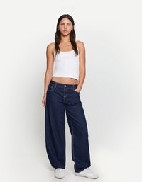 Jean en denim bleu foncé à jambes larges avec des coutures contrastées, dotés de poches avant et d'une fermeture à bouton classique, associés à un débardeur blanc.