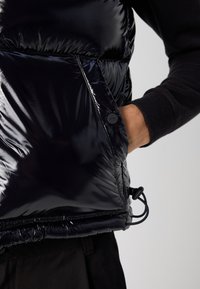 Hand in der Tasche einer glänzenden schwarzen Steppweste mit einem Knopf, der "Hugo" beschriftet ist, getragen über einem schwarzen Langarmoberteil und schwarzen Hosen.