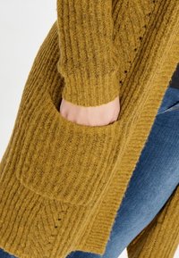 Cardigan a maglione giallo senape con texture a coste, maniche lunghe e tasche frontali. I bordi sono rifiniti con un motivo sottile.