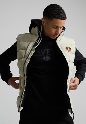 Junger Mann richtet beige, aufgeknöpfte Steppweste mit schwarzem Hoodie darunter, Marke "Zavetti Canada" sichtbar auf Weste und Hoodie.