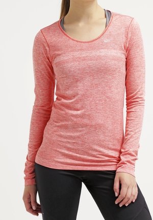 Femme portant un haut de sport à manches longues rose chiné avec des leggings noirs, debout, une main détendue le long du corps.