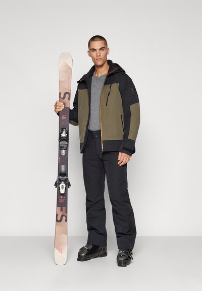 Γεννήτρια σκι με μαύρη και ελαιογραφική ζακέτα, γκρι μπλούζα, μαύρο παντελόνι και παπούτσια σκι, κρατώντας σκι με δεσίματα, μπροστά σε απλό φόντο.