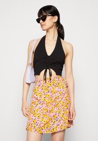 Top cropped preto com alças e detalhe franzido, combinado com uma saia envolvente floral rosa com flores amarelas e rosas. A modelo segura uma bolsa lavanda.