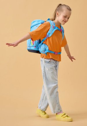 Blauer, gemusterter Rucksack mit pinken Trägern, getragen von einem Kind in einem orangen Hemd und abgenutzten hellen Jeans, kombiniert mit gelben Schuhen.