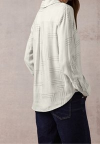 Chemise gris clair à manches longues avec un motif géométrique subtil, poignets à boutons et ourlet incurvé. Portée avec un pantalon bleu foncé.