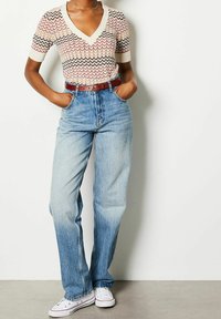 Person wearing a multicolored zigzag short-sleeve top, high-waisted light blue jeans with brown belt, white sneakers, hands in pockets.