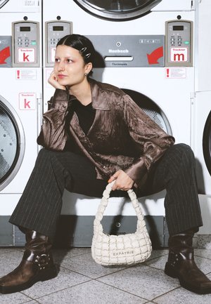 Nuori henkilö, jolla on lyhyet tummat hiukset ja hiusklipsit, istuu pyykin kuivausrumpujen edessä. Hänellä on päällään kiiltävä ruskea paita ja raidalliset housut, ja hän pitää kädessään valkoista laukkua.